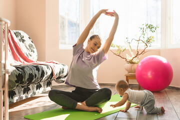 Young happy mother is practicing fitness and yoga with her little child on the sports mat at home. Self-isolation indoor sports