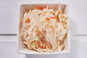 Sauerkraut salad with carrots and spices in a white square porcelain bowl on a white wooden table. Traditional Russian snack.