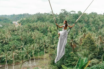 Foto op Canvas Tanned beautiful woman in a long white dress with a train, riding on a swing. In the background, a rainforest and palm trees. Copy space. Rear view © _KUBE_