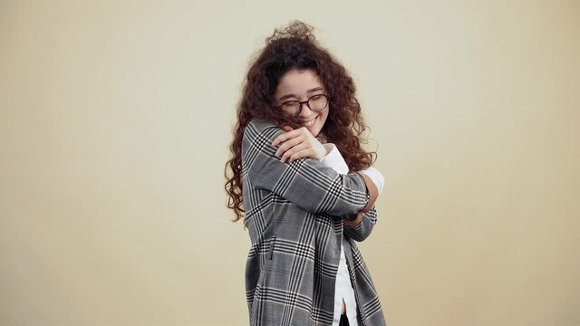 The happy, cheerful young woman with her hands embraces warmly, inspires love and protection. Cretaceous in gray jacket and white shirt, with glasses posing isolated on a beige background in the