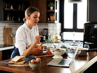 Young woman cooking in the kitchen. Beautiful woman following recipe on laptop and preparing delicious food.