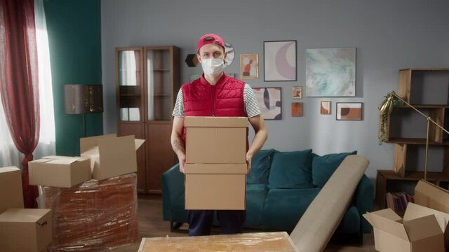 Delivery Man With Covid Mask Holding Carton In New Apartment And Looking At Camera. Mover Carrying All Furniture And Boxes In Room Or Office. Looking At Camera. Daylight