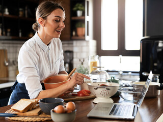 Young woman cooking in the kitchen. Beautiful woman following recipe on laptop and preparing...