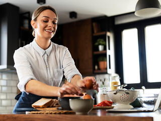 Young woman cooking in the kitchen. Beautiful woman following recipe on laptop and preparing delicious food.