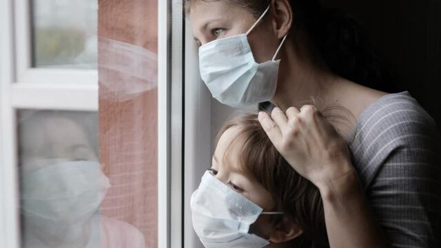 Mature Woman With Her Son, With Protective Mask, Sad Looking Through Window Worried About Covid-19 Lockdown. Family In Protection Mask Looking Out Window Home. Quarantine Coronavirus Social Distancing