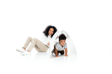 little african american boy crawling under paper roof near smiling mother on white
