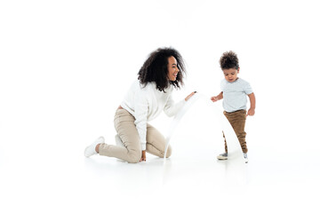 full length view of cheerful african american woman and toddler son near paper roof on white