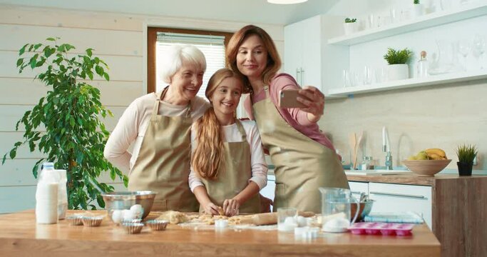 Happy grandmother, granddaughter and mother prepare cookies for guests in kitchen and take photo together on smartphone.