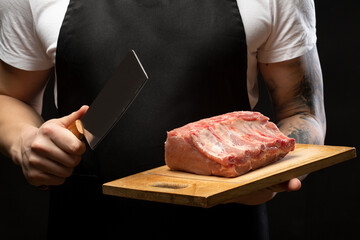 A butcher in a black apron holds pork ribs and a meat hatchet. Close-up.
