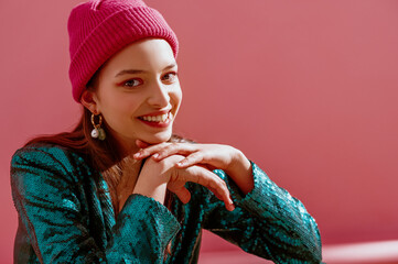 Close up portrait of young beautiful happy smiling fashion model wearing trendy pink, fuchsia color beanie hat, earrings, green sequin blazer. Spring fashion conception. Copy, empty space for text