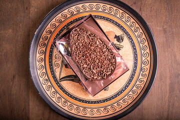 Top view of a stuffed chocolate easter egg with grated chocolate on the top and a spoon on the side in a 
transparent package on a plate with an african design on a wooden table.
