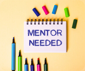 The words MENTOR NEEDED written in a white notebook on a beige background near multi-colored markers