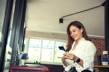 An Asian business woman sits in a coffee shop at a wooden table, drinks coffee and uses a smartphone on the table with a laptop. A woman holding a phone and looking at the screen