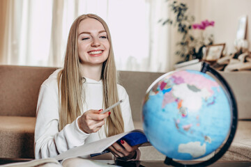 Caucasian student girl smiles, holds a book in her hand and studies geography on a globe while sitting at home in quarantine. Education abroad. Distance learning