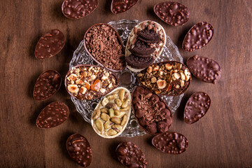 Fototapeta premium Top view of six mini stuffed easter eggs on a glass plate and mini crunchy stuffed chocolate with peanut easter eggs around it on a wooden table.