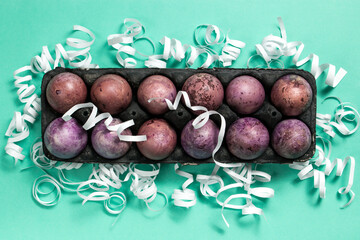 Colorful painted eggs in a black egg box on the turquoise background. White decorations. Easter flat lay with copy space