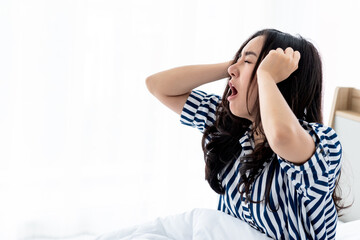 Asian attractive young woman sitting on a clean white bed, She covered her mouth and yawning After she woke up, concept to people and Resting with insufficient sleep Causing drowsiness.