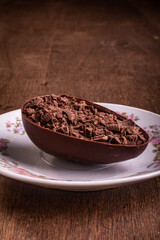 Mini stuffed chocolate easter egg with grated chocolate on the top on a white plate with flowers design on a wooden table with free space at the top.