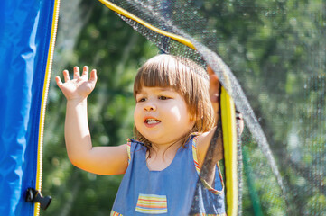 A small, emotional, beautiful girl stands and looks through the net at the trampoline. Cute, active little girl playing outdoors in the playground. Happy childhood. Summer holidays