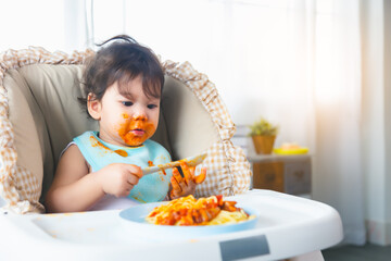 Adorable little toddler girl or infant baby eating delicious food by herself Cute infant child get hungry and eating food by practice using spoon Mix race children get dirty mouth and hand Health care