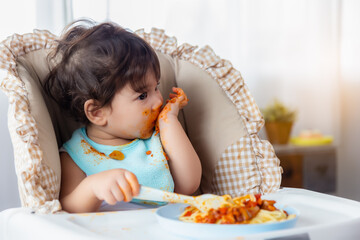 Adorable little toddler girl or infant baby eating delicious food by herself Cute infant girl get hungry and eating food by using hand Mix race daughter get dirty around mouth, hand Child enjoy eating