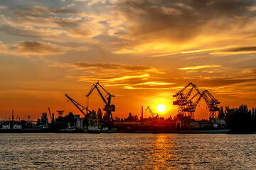 Fototapeta na wymiar Sunset silhouette of Kherson port (Ukraine). The sun sets behind the port cranes against the background of an orange sky and reflections in the water of the Dnieper River