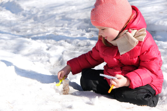 Little girl in warm winter clothes with notepad and pen in her hand investigate details of nature and experimenting with dry plants snow and laboratory flask. Winter outdoor kids activity learning