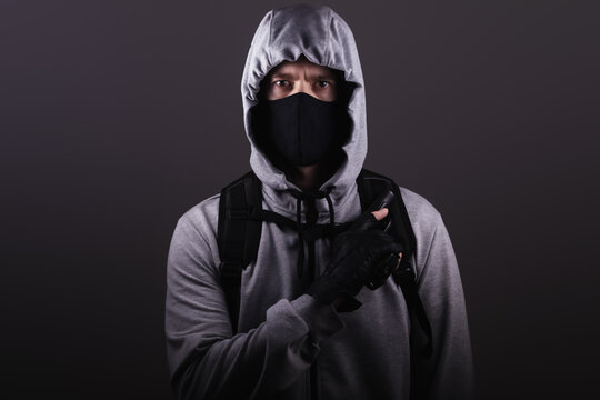 A man in a mask holds a gun in his hand, wearing a hooded jacket over his head, looks at the camera