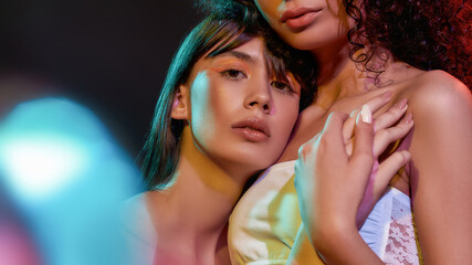 Close up portrait of two artistic young female models with professional art makeup hugging, posing together in neon light isolated over black background