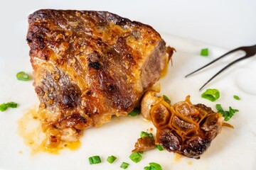 Piece of freshly baked pork neck meat and roasted garlic on white kitchen board, fork.