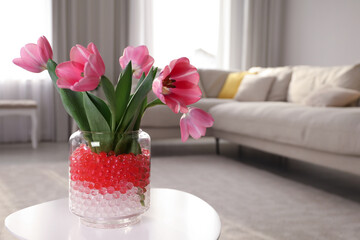 Different color fillers and tulips in glass vase on white table at home, space for text. Water beads