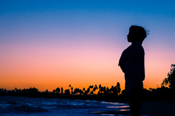 Silhueta de uma criança enrolada numa toalha olhando para o horizonte durante lusco fusco do pôr do sol em uma praia.