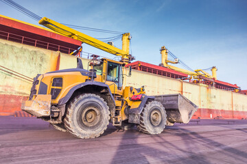 A bucket loader stands on the dock of the port against the backdrop of a cargo ship.