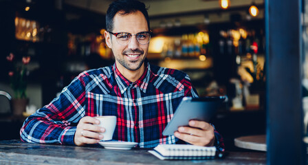 Portrait of smiling man looking at camera during coffee break for updating application on digital tablet, Caucasian male blogger in eyewear connecting to free 4G high speed internet on gadget