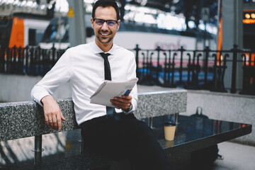 Caucaisan male economist dressed in formal clothes sitting at bench waiting for railway train and smiling at camera, portrait of good looking businessman in optical spectacles holding newspaper