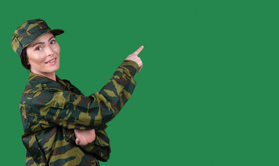 A girl in a military uniform points with a forefinger to an empty space and looks at the camera with a smile. Green background and side space for text or advertisements.