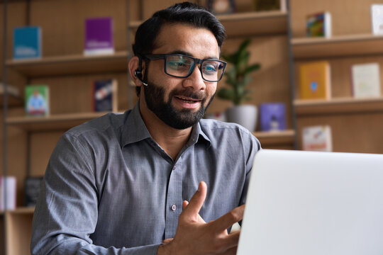 Indian Business Man Speaking Having Virtual Meeting On Laptop. Professional Remote Online Customer Service Support Manager Wearing Headset Talking Consulting On Video Call At Home Office Call Center.