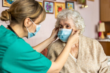 Female nurse helping a senior woman to put on protective face mask during home visit
