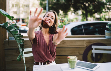 Happy caucasian female in trendy wear sitting on cafe terrace holding sunglasses enjoying free time, smiling woman satisfied with free time on terrace with green smoothie recreating during work break