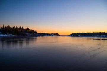 Obraz premium A peaceful winter scenery of frozen seashore with a hut in a chilly weather with clear sky horizon, sunrays and pine trees