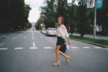 Fototapeta na wymiar Full length portrait of cheerful female tourist dressed in trendy wear crossing pedestrian road during travel vacations, happy Caucasian woman smiling at camera while walking in urban city
