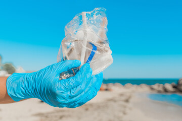 The hands of a man in blue household protective gloves hold a pile of garbage in a bag against the sky and sea beach, close-up