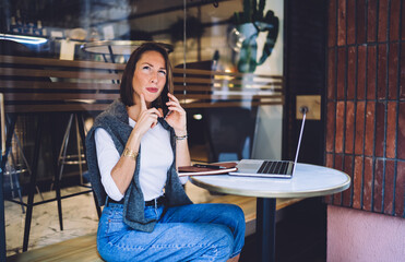 Young caucasian woman in casual wear making smartphone call during remote job in cafeteria, serious female freelancer pondering on delivery talking on mobile phone on remote job with service operator