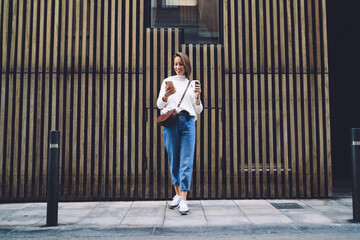 Confident woman standing near modern building with smartphone and coffee