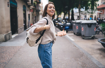 Cheerful female tourist with travel backpack standing at urban street and smiling satisfied with...