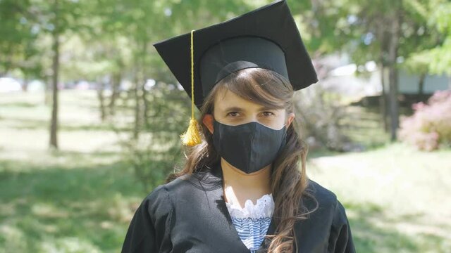 A female graduate student in a graduation gown removes a medical mask during the coronavirus pandemic.