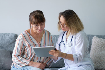 Female professional doctor showing medical test result explaining prescription using digital tablet app visiting senior woman patient at home sitting on sofa. Elderly people healthcare tech concept.