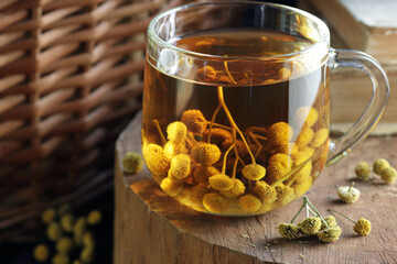 Tansy seed tea or infusion in a glass cup with flowers on wood with books nearby, herbal drink is...