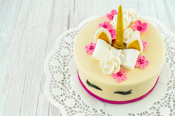 Unicorn cake on a wooden background and lace. Cute unicorn layered homemade cake decorated with bow, rose flowers, golden corn and ears. Copy space