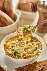 Soup with noodle, pieces of chicken and vegetables in white bowl served with spices and breads on light wooden background.